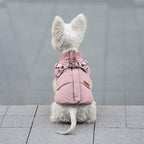 Rear view of a small white dog wearing a pink padded harness vest, sitting on a gray paved surface.