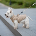 Small white dog wearing a beige padded harness vest on a leash, standing on concrete steps outdoors.